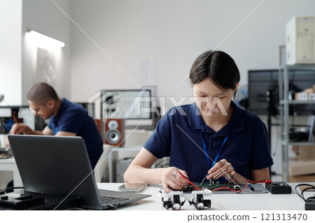 Asian engineer fixing circuit board while seated in modern lab. Background includes colleague working and various electronic equipment present 121313470