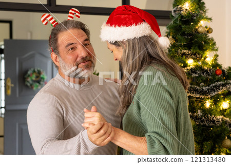 Mature couple dancing joyfully by Christmas tree, wearing festive holiday hats, at home 121313480
