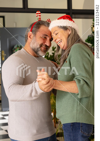 Christmas time, Joyful mature couple dancing at home, wearing festive holiday hats, smiling warmly 121313482