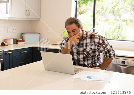 Mature man working on laptop in kitchen, focusing on online tasks, at home 121313576