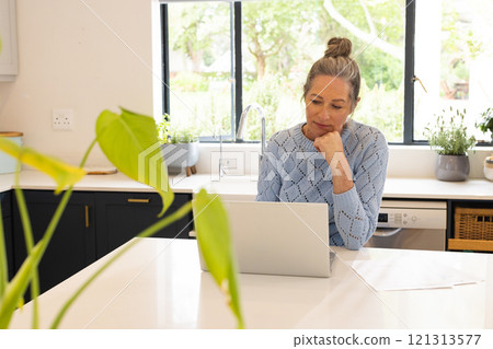 Mature woman working on laptop in bright kitchen, focused and thoughtful, at home 121313577