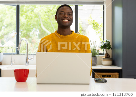 Smiling African american man in yellow sweater using laptop at home with red mug nearby 121313700