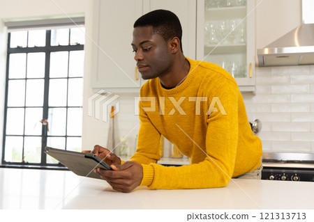 African american man in kitchen using tablet, wearing yellow sweater, focused on screen, at home African american man in kitchen using tablet, wearing yellow sweater, focused on screen, at home 121313713