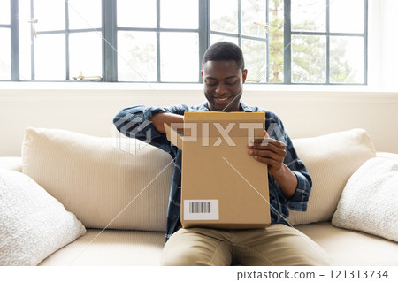 Smiling African american man opening package on couch at home, enjoying holiday surprise Smiling African american man opening package on couch at home, enjoying holiday surprise 121313734