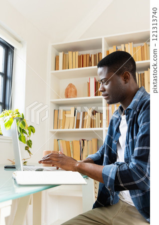 Focused African american man working on laptop at home office with bookshelves behind 121313740