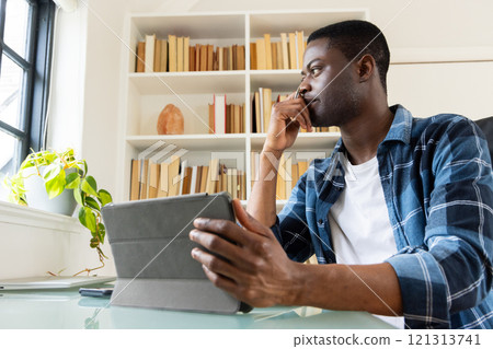 African american man using tablet at home, thinking while sitting by window and bookshelf 121313741