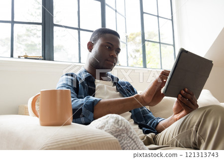 Relaxing at home, African american man using tablet with coffee mug on cozy couch Relaxing at home, African american man using tablet with coffee mug on cozy couch 121313743