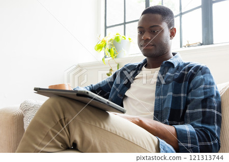 Relaxing at home, young African american man using tablet with coffee on cozy sofa Relaxing at home, young African american man using tablet with coffee on cozy sofa 121313744