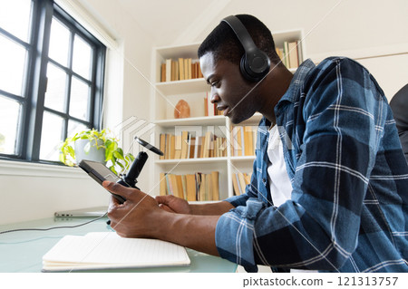 African american man wearing headphones recording podcast at home using smartphone and microphone 121313757