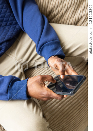African American man using smartphone on couch, wearing blue sweater, at home African American man using smartphone on couch, wearing blue sweater, at home 121313803
