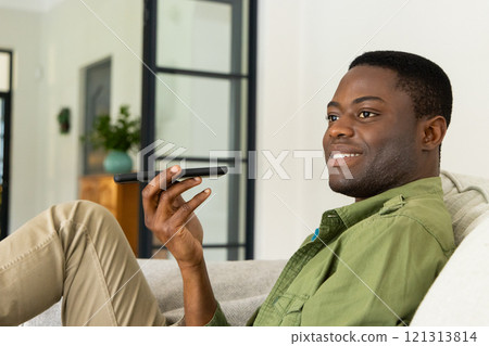 Relaxing at home, African american man using smartphone for voice message, smiling contentedly 121313814