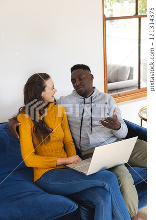 Happy diverse couple sitting on sofa using laptop at home 121314375