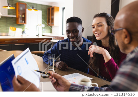 Diverse couple and biracial male financial advisor using laptop and discussing documents at home 121314530