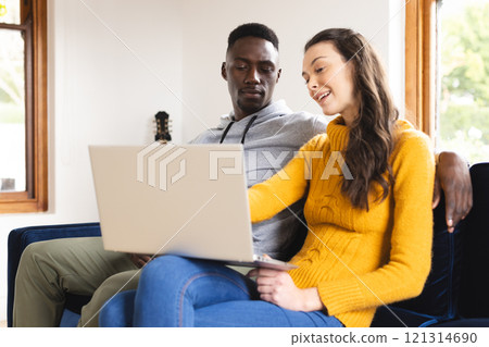 Happy diverse couple sitting on sofa using laptop at home 121314690