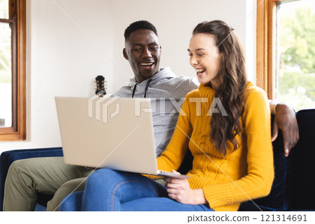 Happy diverse couple sitting on sofa using laptop at home 121314691