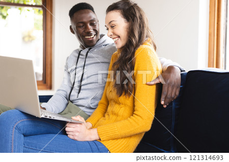 Happy diverse couple sitting on sofa using laptop at home 121314693