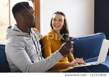 Happy diverse couple sitting on sofa using laptop for online shopping at home 121314698