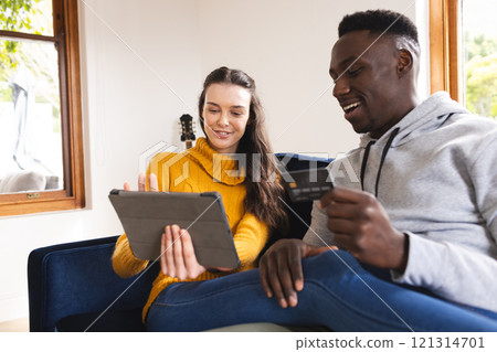 Happy diverse couple sitting on sofa using tablet for online shopping at home 121314701