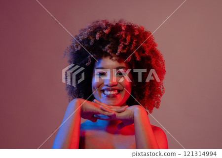Smiling biracial woman wearing pink eyeshadow and lipstick in blue and red light, hands under chin Smiling biracial woman wearing pink eyeshadow and lipstick in blue and red light, hands under chin 121314994