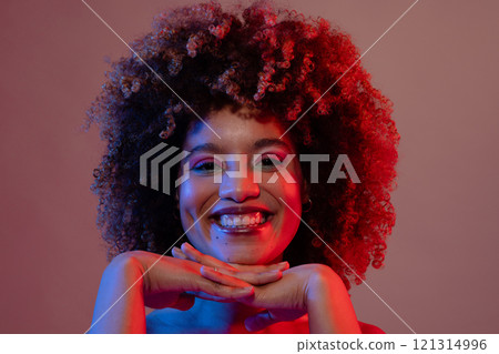 Smiling biracial woman wearing pink eyeshadow and lipstick in blue and red light, hands under chin 121314996