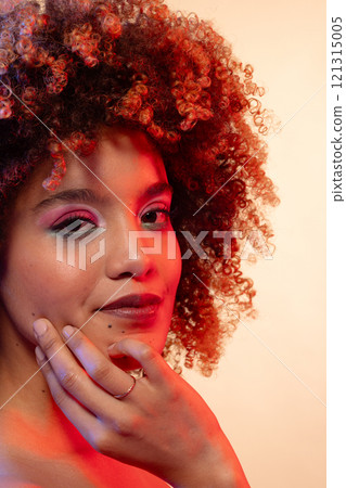 Happy biracial woman with curly hair in pink eyeshadow and lipstick touching face in red light Happy biracial woman with curly hair in pink eyeshadow and lipstick touching face in red light 121315005