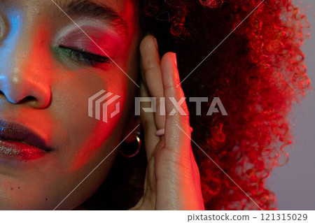 Biracial woman wearing eyeshadow and lipstick touching head with eyes closed in red and blue light 121315029