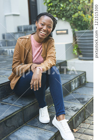 Portrait of happy african american woman wearing leather jacket sitting on stairs outside house 121315300