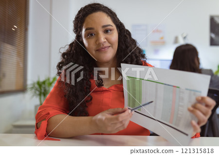 Smiling biracial businesswoman sitting at desk making video call holding documents in modern office Smiling biracial businesswoman sitting at desk making video call holding documents in modern office 121315818
