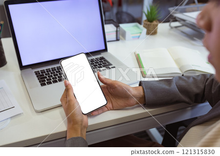 Biracial businesswoman sitting at desk using laptop and smartphone with copy space in modern office Biracial businesswoman sitting at desk using laptop and smartphone with copy space in modern office 121315830