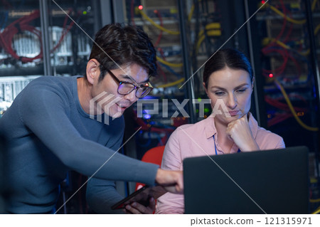 Diverse male and female engineers discussing over a laptop in computer server room 121315971