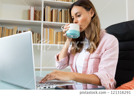Woman working on laptop at home, sipping coffee, surrounded by books 121316291