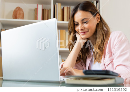 At home, woman using laptop and smiling, enjoying online holiday shopping 121316292