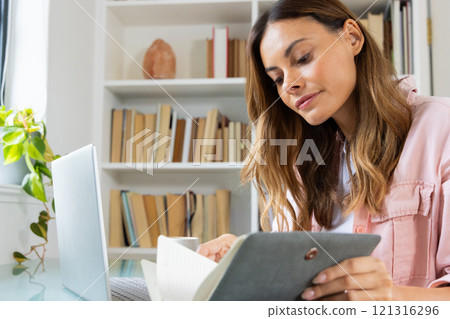 At home, woman reading notebook and using laptop, focusing on work At home, woman reading notebook and using laptop, focusing on work 121316296