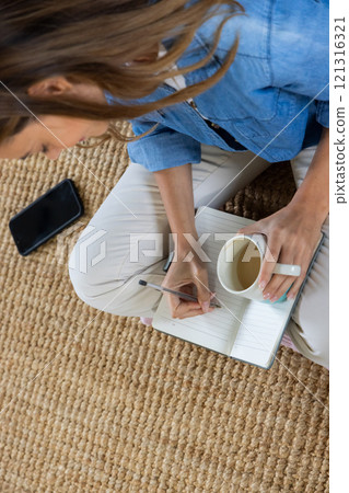 Writing in notebook, woman holding coffee cup, sitting on woven rug at home Writing in notebook, woman holding coffee cup, sitting on woven rug at home 121316321