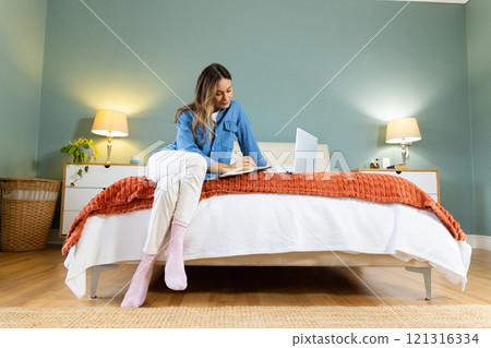 Woman sitting on bed writing in notebook with laptop, cozy bedroom setting, at home Woman sitting on bed writing in notebook with laptop, cozy bedroom setting, at home 121316334