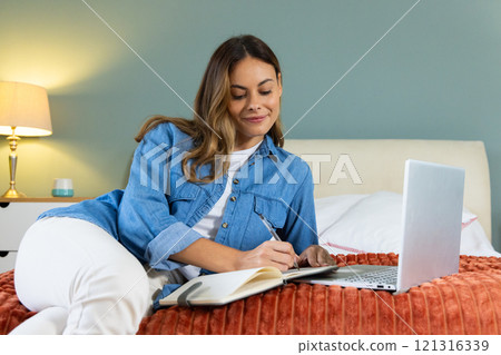 At home, woman writing in notebook while using laptop on bed, feeling relaxed 121316339