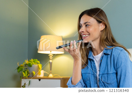 Smiling woman using smartphone voice assistant at home, relaxing on couch 121316340