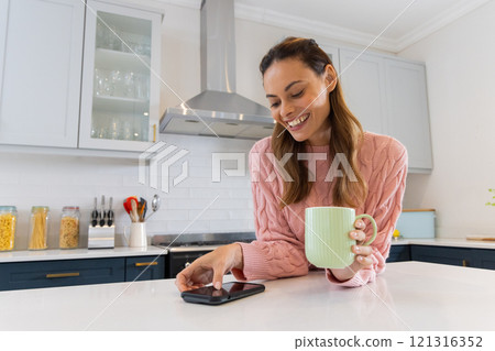 Smiling woman in kitchen using smartphone and holding coffee mug, at home 121316352