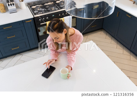 Woman in kitchen using smartphone and holding coffee mug, enjoying morning routine, at home Woman in kitchen using smartphone and holding coffee mug, enjoying morning routine, at home 121316356