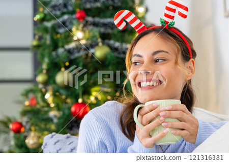 Smiling woman with festive headband enjoying hot drink by Christmas tree, at home Smiling woman with festive headband enjoying hot drink by Christmas tree, at home 121316381