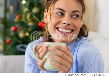 Smiling woman enjoying hot drink at home near Christmas tree 121316385