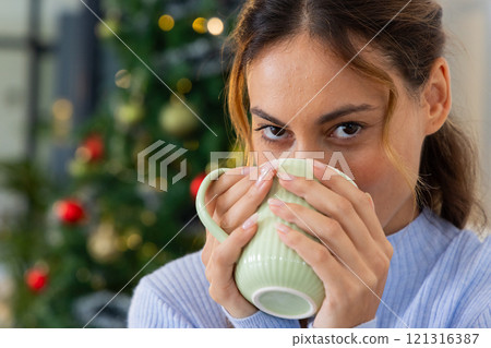 Woman enjoying hot drink by Christmas tree, feeling cozy and festive, at home 121316387