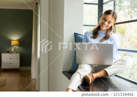 Smiling woman using laptop by window at home, enjoying cozy atmosphere Smiling woman using laptop by window at home, enjoying cozy atmosphere 121316402