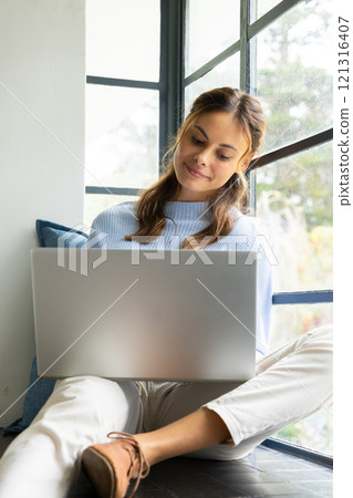 Smiling woman using laptop by window, enjoying cozy home atmosphere 121316407