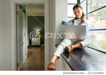 Woman sitting by window using laptop and holding credit card, shopping online, at home 121316408