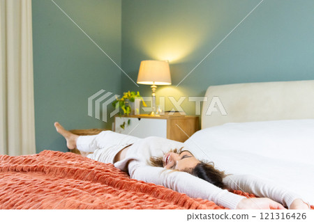 Relaxing on bed, woman stretching joyfully in cozy bedroom with warm lighting, at home 121316426