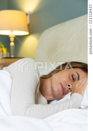 Woman peacefully sleeping in cozy bedroom with warm lamp light, at home 121316437
