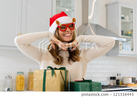 Smiling woman in Santa hat posing with Christmas gifts in kitchen, at home Smiling woman in Santa hat posing with Christmas gifts in kitchen, at home 121316443