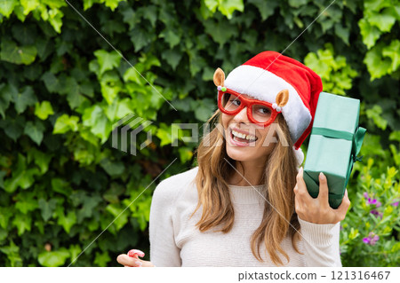 Smiling woman in Santa hat holding gift outdoors, celebrating Christmas joy, copy space Smiling woman in Santa hat holding gift outdoors, celebrating Christmas joy, copy space 121316467