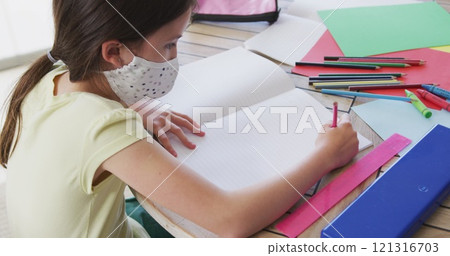 Caucasian girl spending time at home wearing face mask, sitting at table doing her homework, in slow 121316703